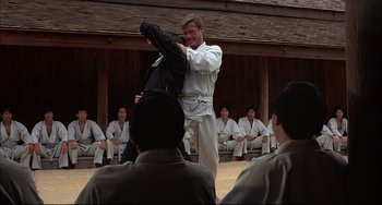 Movie still from “The Man with the Golden Gun” (1974), directed by Guy Hamilton – A man in a white shirt and white pants is holding a black shirt; Medium shot, Over the shoulder angle