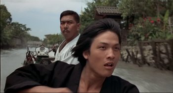 Movie still from “The Man with the Golden Gun” (1974), directed by Guy Hamilton – Two men riding motorcycles down a street in a village; Close Up shot, Low angle