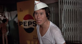 Movie still from “The Man with the Golden Gun” (1974), directed by Guy Hamilton – A man wearing a paper hat in front of a pepsi sign; Close Up shot, Low angle