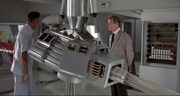 Movie still from “The Man with the Golden Gun” (1974), directed by Guy Hamilton – A man standing next to a machine in a room; Wide shot, High angle