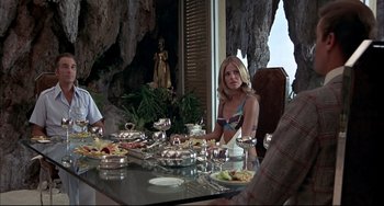 Movie still from “The Man with the Golden Gun” (1974), directed by Guy Hamilton – A group of people sitting at a table with plates of food on top of it; Medium shot, Low angle