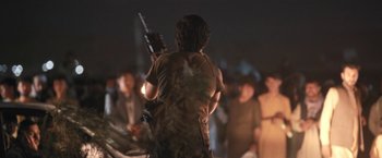 Movie still from “Retrograde” (2022), directed by Matthew Heineman – A man holding a gun while standing in front of a group of people; Medium shot, Over the shoulder angle