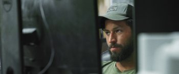Movie still from “Retrograde” (2022), directed by Matthew Heineman – A man with a beard wearing a hat looking at a computer screen; Close Up shot, Over the shoulder angle