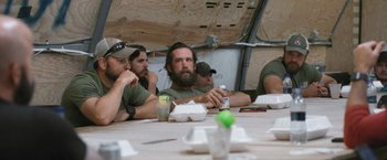 Movie still from “Retrograde” (2022), directed by Matthew Heineman – A group of men sitting at a table with food; Medium shot, Over the shoulder angle