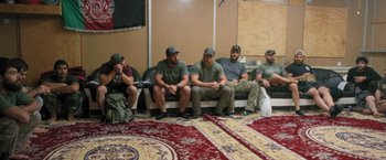 Movie still from “Retrograde” (2022), directed by Matthew Heineman – A group of men sitting on a couch in a room; Wide shot, Over the shoulder angle