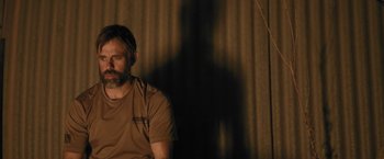 Movie still from “Retrograde” (2022), directed by Matthew Heineman – A man sitting in front of a curtain with his shadow on the wall; Medium shot, Over the shoulder angle
