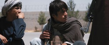 Movie still from “Retrograde” (2022), directed by Matthew Heineman – A young man holding a gun while sitting on a chair; Close Up shot, Low angle