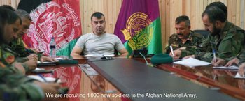 Movie still from “Retrograde” (2022), directed by Matthew Heineman – A man sitting at a table in front of an afghan national flag; Medium shot, Over the shoulder angle