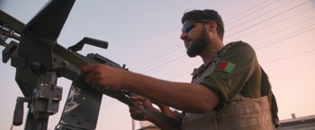 Movie still from “Retrograde” (2022), directed by Matthew Heineman – A man holding a gun while standing next to a pole; Medium shot, Low angle
