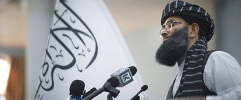 Movie still from “Retrograde” (2022), directed by Matthew Heineman – A man with a long beard talking into microphones in front of a wall; Close Up shot, Low angle