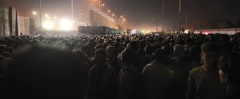 Movie still from “Retrograde” (2022), directed by Matthew Heineman – A crowd of people standing in the dark at night; Extreme Wide shot, High angle