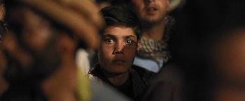 Movie still from “Retrograde” (2022), directed by Matthew Heineman – A young man looking into the camera while sitting in a crowd; Close Up shot, High angle