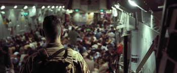 Movie still from “Retrograde” (2022), directed by Matthew Heineman – A man with a back pack standing in front of a crowd of people; Medium shot, Over the shoulder angle