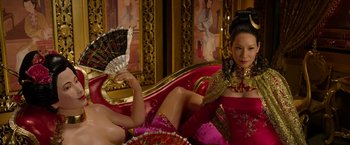 Movie still from “The Man with the Iron Fists” (2012), directed by RZA – A woman sitting on top of a couch next to another woman holding a fan; Medium shot, Over the shoulder angle