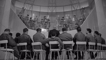 Movie still from “The Manchurian Candidate” (1962), directed by John Frankenheimer – A black and white photo of a group of people sitting in chairs; Wide shot, High angle
