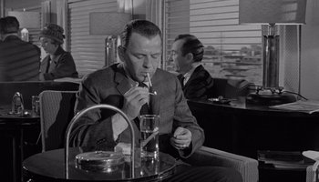 Movie still from “The Manchurian Candidate” (1962), directed by John Frankenheimer – A man smoking a cigarette while sitting at a table in front of another man; Medium shot, Over the shoulder angle