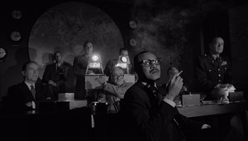 Movie still from “The Manchurian Candidate” (1962), directed by John Frankenheimer – Black and white photograph of a man smoking a cigarette in front of a group of people; Medium shot, Low angle