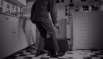 Movie still from “The Manchurian Candidate” (1962), directed by John Frankenheimer – A man in a suit is standing in a kitchen with a suitcase; Wide shot, Low angle