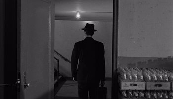 Movie still from “The Manchurian Candidate” (1962), directed by John Frankenheimer – A black and white photo of a man in a suit and hat holding a briefcase; Wide shot, Over the shoulder angle