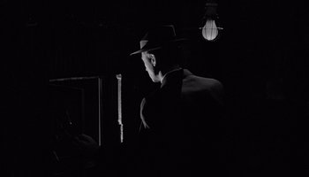 Movie still from “The Manchurian Candidate” (1962), directed by John Frankenheimer – A man wearing a hat and a suit is standing in a dark room; Medium shot, Low angle