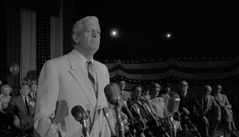 Movie still from “The Manchurian Candidate” (1962), directed by John Frankenheimer – An older man standing in front of microphones in front of a group of people; Medium shot, Low angle