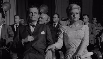 Movie still from “The Manchurian Candidate” (1962), directed by John Frankenheimer – An older man and woman sitting next to each other in front of other people; Medium shot, Low angle