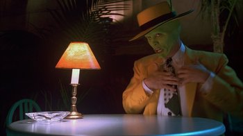 Movie still from “The Mask” (1994), directed by Chuck Russell – A man in a yellow suit and yellow hat standing in front of a table with a lamp; Medium shot, Over the shoulder angle