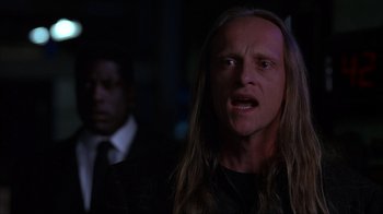 Movie still from “The Mask” (1994), directed by Chuck Russell – A man with long blonde hair and a black shirt; Close Up shot, Over the shoulder angle