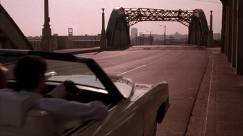 Movie still from “The Mask” (1994), directed by Chuck Russell – A car driving down a street with a bridge in the background; Extreme Wide shot, Over the shoulder angle