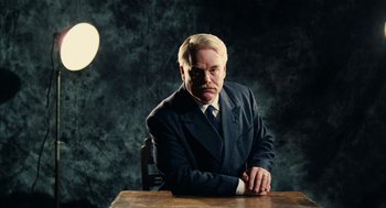 Movie still from “The Master” (2012), directed by Paul Thomas Anderson – A man sitting at a wooden table in a suit; Medium shot, Low angle