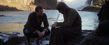 Movie still from “The Mission” (1986), directed by Roland Joffé – Two men sitting next to a body of water; Medium shot, Over the shoulder angle