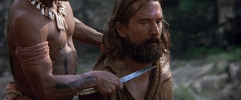 Movie still from “The Mission” (1986), directed by Roland Joffé – A man with long hair and a beard cutting something with a knife; Close Up shot, Over the shoulder angle