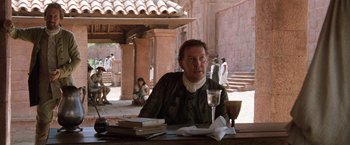 Movie still from “The Mission” (1986), directed by Roland Joffé – A man sitting at a table with a glass of wine; Medium shot, Over the shoulder angle