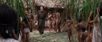 Movie still from “The Mission” (1986), directed by Roland Joffé – A group of people standing around a man wearing a black robe; Wide shot, Over the shoulder angle