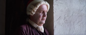 Movie still from “The Mission” (1986), directed by Roland Joffé – An old man wearing a wig and a wig stand in front of a wall; Close Up shot, Low angle