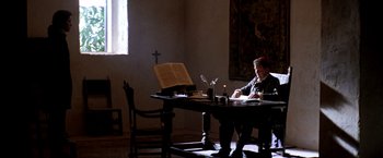 Movie still from “The Mission” (1986), directed by Roland Joffé – A person sitting at a table with a book and a candle; Wide shot, Low angle