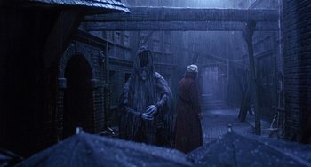 Movie still from “The Muppet Christmas Carol” (1992), directed by Brian Henson – A man in a hooded cloak and a woman in a red coat in the rain with an umbrella; Wide shot, Over the shoulder angle