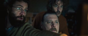 Movie still from “Riders of Justice” (2020), directed by Anders Thomas Jensen – A man with a beard and mustache looking at the camera with two other men in the background; Close Up shot, Low angle