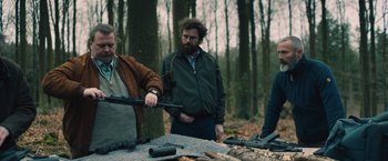 Movie still from “Riders of Justice” (2020), directed by Anders Thomas Jensen – A man holding a gun while standing next to another man in the woods; Medium shot, Over the shoulder angle