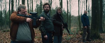 Movie still from “Riders of Justice” (2020), directed by Anders Thomas Jensen – A man holding a gun while standing next to another man holding a rifle; Medium shot, Low angle