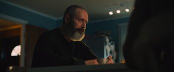 Movie still from “Riders of Justice” (2020), directed by Anders Thomas Jensen – A man with a beard is sitting at a table with a cup of coffee; Medium shot, Low angle