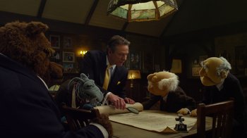 Movie still from “The Muppets” (2011), directed by James Bobin – A man in a suit and tie cutting a sheet of parchment paper with a stuffed animal; Medium shot, Low angle