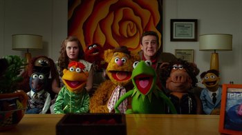 Movie still from “The Muppets” (2011), directed by James Bobin – A group of people sitting in front of a painting of kermit the frog and fozzie the bear; Medium shot, Over the shoulder angle