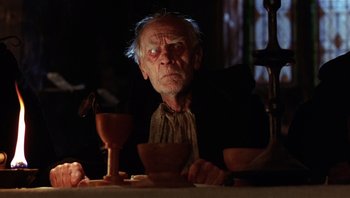 Movie still from “The Name of the Rose” (1986), directed by Jean-Jacques Annaud – An old man sitting at a table with a wine glass; Close Up shot, Low angle