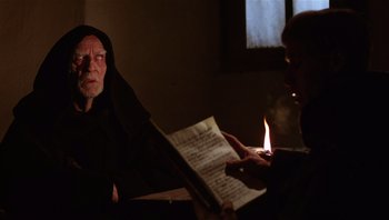 Movie still from “The Name of the Rose” (1986), directed by Jean-Jacques Annaud – An old man sitting at a table reading a book; Medium shot, Over the shoulder angle