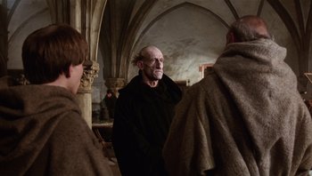Movie still from “The Name of the Rose” (1986), directed by Jean-Jacques Annaud – An old man with a bald head is standing next to two other men; Medium shot, Over the shoulder angle