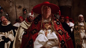 Movie still from “The Name of the Rose” (1986), directed by Jean-Jacques Annaud – An old man wearing a red hat and a white robe; Medium shot, Low angle
