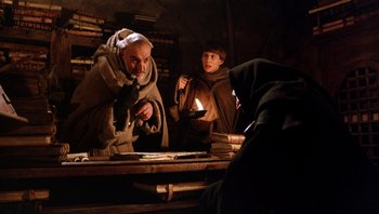 Movie still from “The Name of the Rose” (1986), directed by Jean-Jacques Annaud – Two men are sitting at a table with a candle in the dark; Medium shot, Low angle