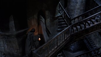 Movie still from “The Name of the Rose” (1986), directed by Jean-Jacques Annaud – A man is standing next to a staircase with a candle; Wide shot, High angle