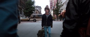 Movie still from “The NeverEnding Story” (1984), directed by Wolfgang Petersen – A young boy walking down the street holding a suitcase; Medium shot, Over the shoulder angle